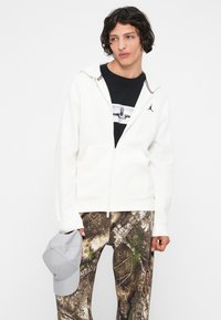 Giovane uomo con una felpa bianca con zip sopra una maglietta nera con grafiche e pantaloni mimetici, che tiene un cappellino da baseball grigio chiaro nella mano sinistra.