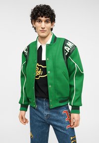 Πράσινο varsity μπουφάν με μαύρα δερμάτινα στοιχεία, κουμπιά snap και λευκή διακόσμηση. Φορεμένο πάνω από ένα μαύρο πουκάμισο με γραφιστικό σχέδιο.