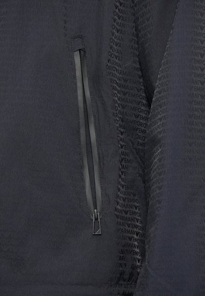 Čierna bunda s textúrovanou látkou. Má vrecko so zipsom na ľavej strane a jemný opakujúci sa vzor loga po celej ploche.