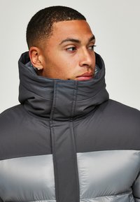 Donkergrijze pufferjack met een glanzende zilveren horizontale strook, hoge kraag en gewatteerde textuur. Heeft stevige stiksels en een slank ontwerp.