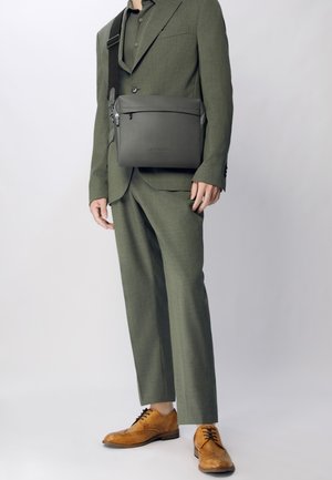 Grå læder messenger taske med tekstureret overflade og lynlås lukning, båret crossbody over et grønt jakkesæt og brune snørebåndssko.