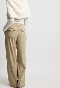 Kvinna i beige vida byxor, vit långärmad tröja och en krämfärgad stickad tröja som hänger över axlarna, står med händerna i fickorna.