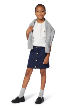 Un cardigan en maille gris drapé sur une chemise blanche avec un logo doré, associé à une jupe navy dotée de boutons dorés et des chaussures noires. Chaussettes grises visibles.