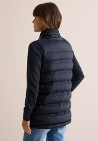 Marinefarbener Steppwesten mit horizontalem Steppmuster. Verfügt über einen hohen Kragen und seitliche Taschen, kombiniert mit einem schwarzen Langarmtop und blauen Jeans.