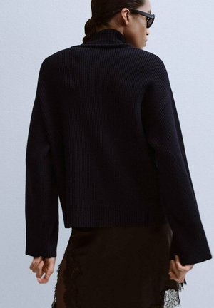 Pull noir à col roulé côtelé avec manches longues, offrant une coupe décontractée et un tissu texturé ; superposé sur une jupe en dentelle noire transparente.