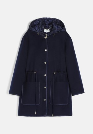 Femme portant un manteau à capuche bleu marine, un pull à col roulé côtelé jaune moutarde et un pantalon en velours côtelé marron avec une ceinture en cuir, posant avec une main dans la poche.