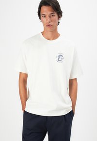 Junger Mann mit dunklen Haaren, der ein weißes, lässig geschnittenes T-Shirt mit einem kleinen blauen Logo "Los Angeles Track & Field" auf der Brust und dunklen Hosen trägt.