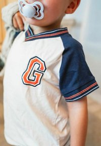 Lyhythihainen paita, jossa valkoinen alaosa ja tummansiniset hihat. Paidan rintamuksessa on brodeerattu oranssi "G"-kirjain ja raidallisia yksityiskohtia.