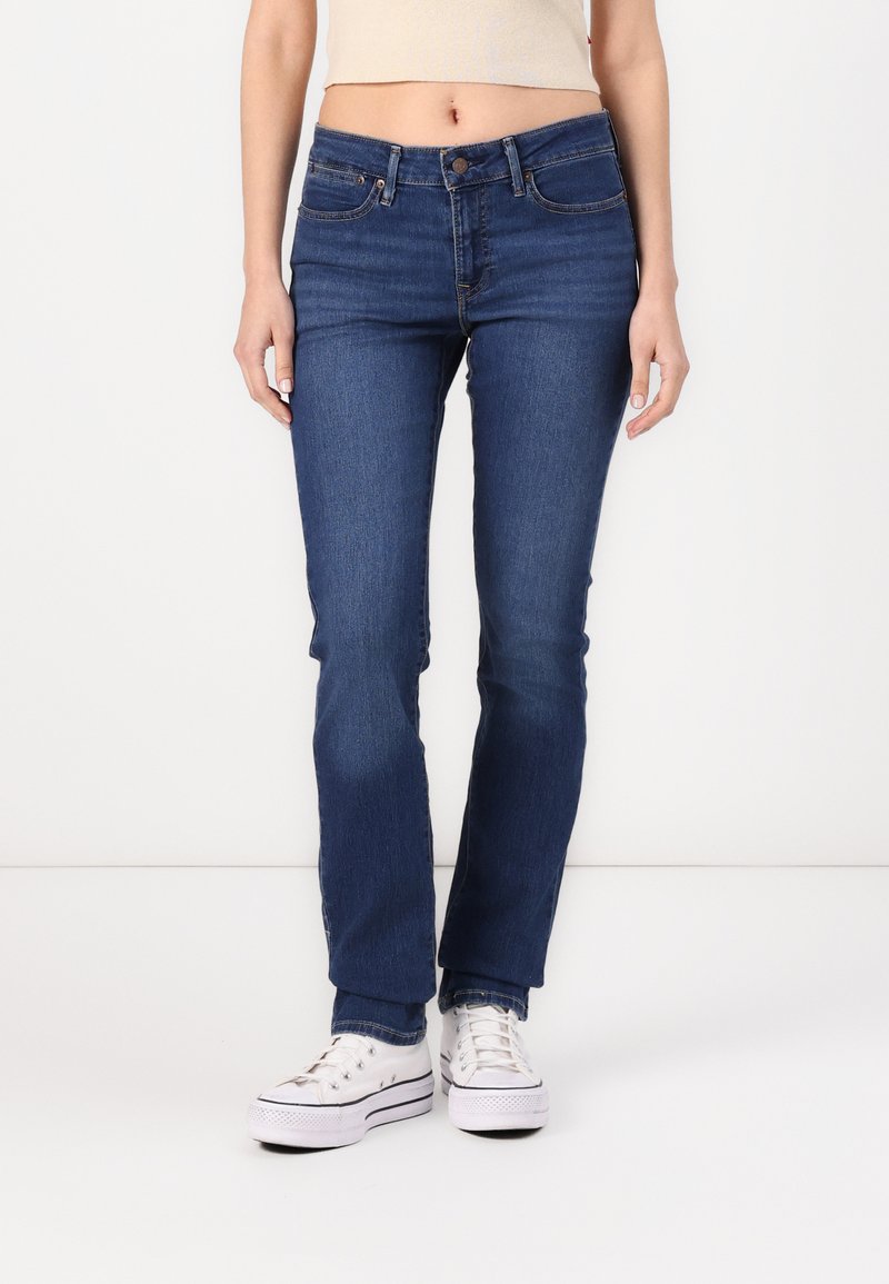 Jeans de denim azul con un corte ajustado, cinco bolsillos y una cintura de altura media, combinados con zapatillas blancas. La tela tiene una textura suave.