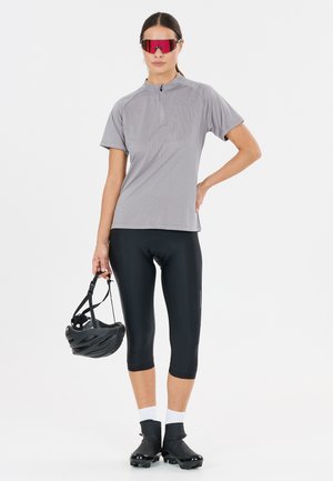 Frau trägt graues Kurzarm-Radtrikot, schwarze Radlerhose, schwarze Radschuhe, rote Sport-Sonnenbrille und hält einen schwarzen Helm.