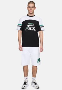 Svart och vit t-shirt med kort ärm och gröna ränder samt FILA-logotyp; matchad med vita shorts som visar en grön FILA-logotyp.