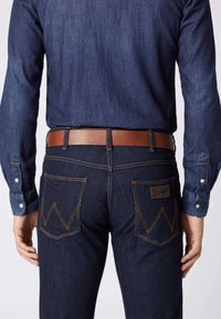 Dunkelblaue Denim-Jeans mit einem Ledergürtel. Merkmale sind sichtbare Nähte und zwei Gesäßtaschen mit einem markanten Muster.