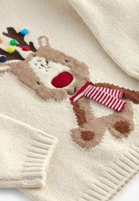 Pull en tricot couleur écrue avec un motif de renne brun, un nez rouge, une écharpe rayée et des pompons colorés sur les bois.