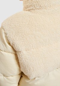Ljusbeige puffjacka med en texturerad, plyschfuskpälskrage. Jackan har en slät tygkropp och mjuka, fluffiga detaljer.