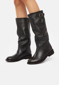 Black leather knee-high boots with a slightly slouched design, prominent stitching, and a buckle accent on the collar. Flat wooden sole.