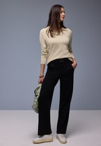 Femme portant un haut beige à manches longues, un pantalon large noir, des baskets blanches, tenant un sac matelassé vert clair, debout devant un fond gris.