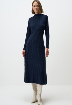Abito a coste blu navy con scollatura alta e maniche lunghe, caratterizzato da un design a zigzag e una silhouette aderente. Stivali bianchi fino al ginocchio.