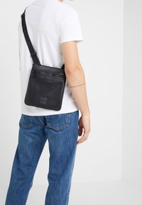 Černá crossbody taška vyrobená z texturované kůže, s vzorem herringbone a kapsami na zip, nošena přes bílé tričko.