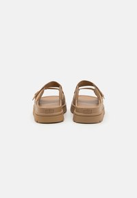 Sandales beiges avec un dos ouvert, une sangle ajustable et une semelle épaisse. Fabriquées en matériau durable, elles présentent une tige texturée et un détail de logo.