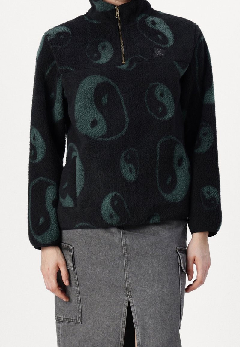 Pull polaire noir avec motifs yin-yang verts, col zippé au quart, porté avec une jupe en jean grise avec une fente devant et des poches latérales.