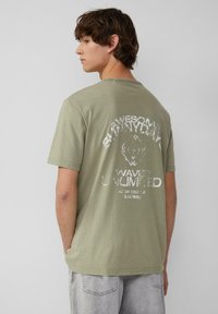 Oliwkowa bawełniana koszulka z krótkim rękawem, z białym nadrukiem na plecach, który zawiera tekst oraz wzór lwa.