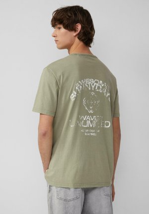 T-shirt en coton vert olive à manches courtes, avec un graphique blanc imprimé au dos comprenant du texte et un design de lion.