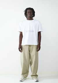 Biała koszulka z krótkim rękawem z jasnoszarym wzorem graficznym, połączona z luźnymi beżowymi spodniami i jasnymi sneakersami. Prosty i schludny wygląd.