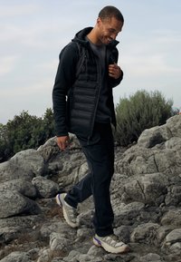 Juodas pūkuotas liemenė virš pilkos palaidinės, derinamas su tamsiomis kelnėmis ir šviesiais žygio bateliais su violetiniais akcentais, vaikščiojant akmenuotu teritorijoje.