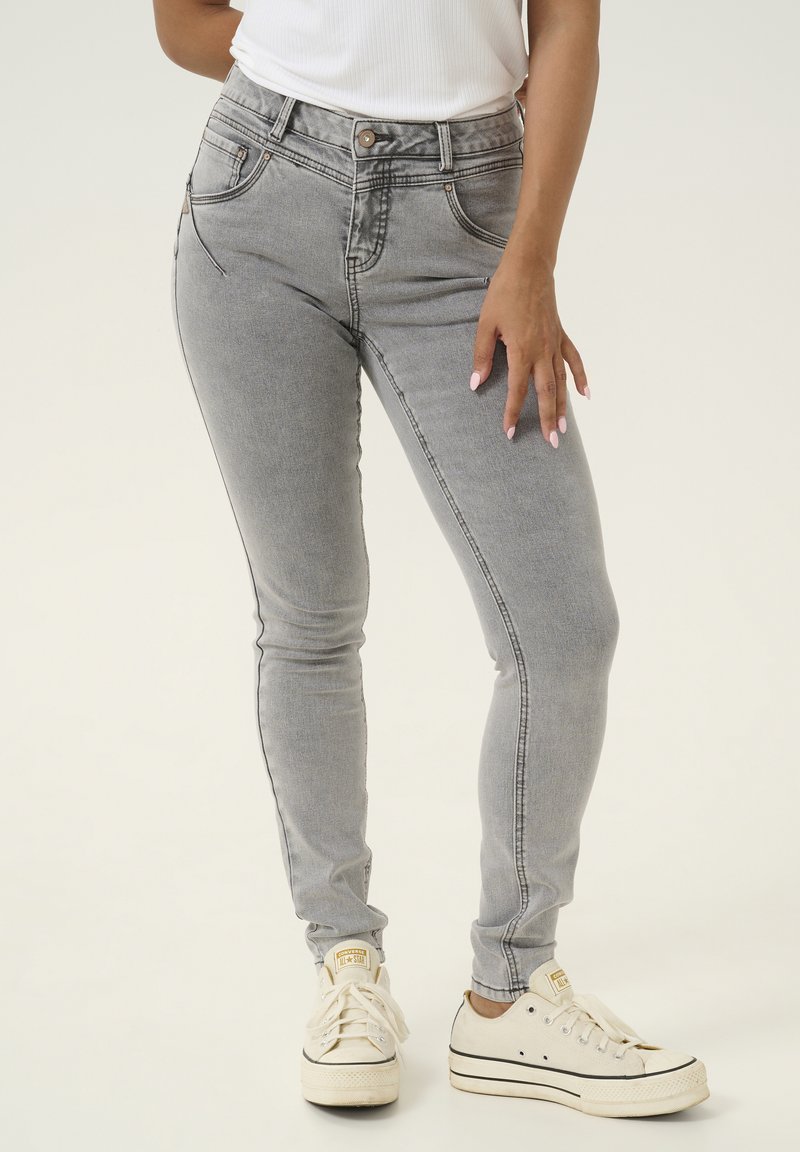 Szürke szűk farmernadrág sima denim textúrával, magas derékkal, elülső zsebekkel és kontrasztos varrással. Fehér sportcipővel párosítva.