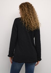 Svart långärmad topp med texturerad tyg, volangkant på fåll och ärmslut, samt en avslappnad passform. Bärs med blå jeans.