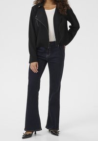 Černá zipová bunda s krátkým střihem, doplněná tmavě modrými rozšířenými džíny a černými lakovanými podpatky. Oblečení zdůrazňuje klasickou siluetu.