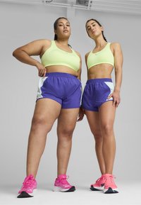 Două femei stau una lângă cealaltă. Una poartă un top scurt neon galben și pantaloni scurți purpurii; cealaltă poartă o ținută similară cu accente bleu deschis. Ambele au adidași roz.