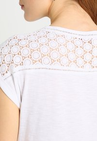 Top blanco con un diseño de encaje en la parte posterior con patrones circulares, mangas cortas y una tela suave y texturizada.