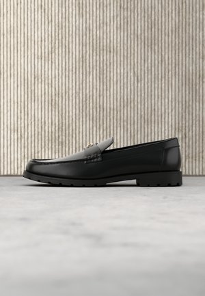Sort læderloafer med chunky sål og guldspænde, placeret på en grå overflade med en struktureret stribet væg i baggrunden.