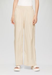 Beige, lichtgewicht, wijdvallende broek met een gestructureerd verticaal streepjespatroon, elastische tailleband en zijzakken. Gecombineerd met witte platformschoenen.