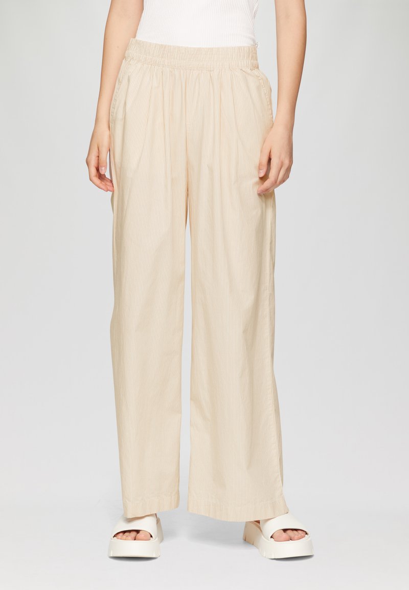 Beige, lichtgewicht, wijdvallende broek met een gestructureerd verticaal streepjespatroon, elastische tailleband en zijzakken. Gecombineerd met witte platformschoenen.