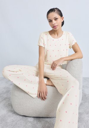 Kremfarvet lounge-sæt med ribstrik og lyserøde hjerte mønstre, med bølget kant. Løst fit top og bukser med vidde ben, båret mens sidder.