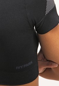 Primer plano del brazo superior de una persona que lleva una camiseta deportiva de manga corta negra con detalles de malla y el logo "IVY PARK" en el lateral.