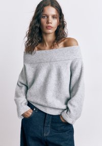 Mladá žena s kudrnatými vlasy, oblečená v šedém svetru se spuštěnými rameny a tmavě modrých džínách, s rukama v kapsách, čelem k přední straně na jednobarevném pozadí.