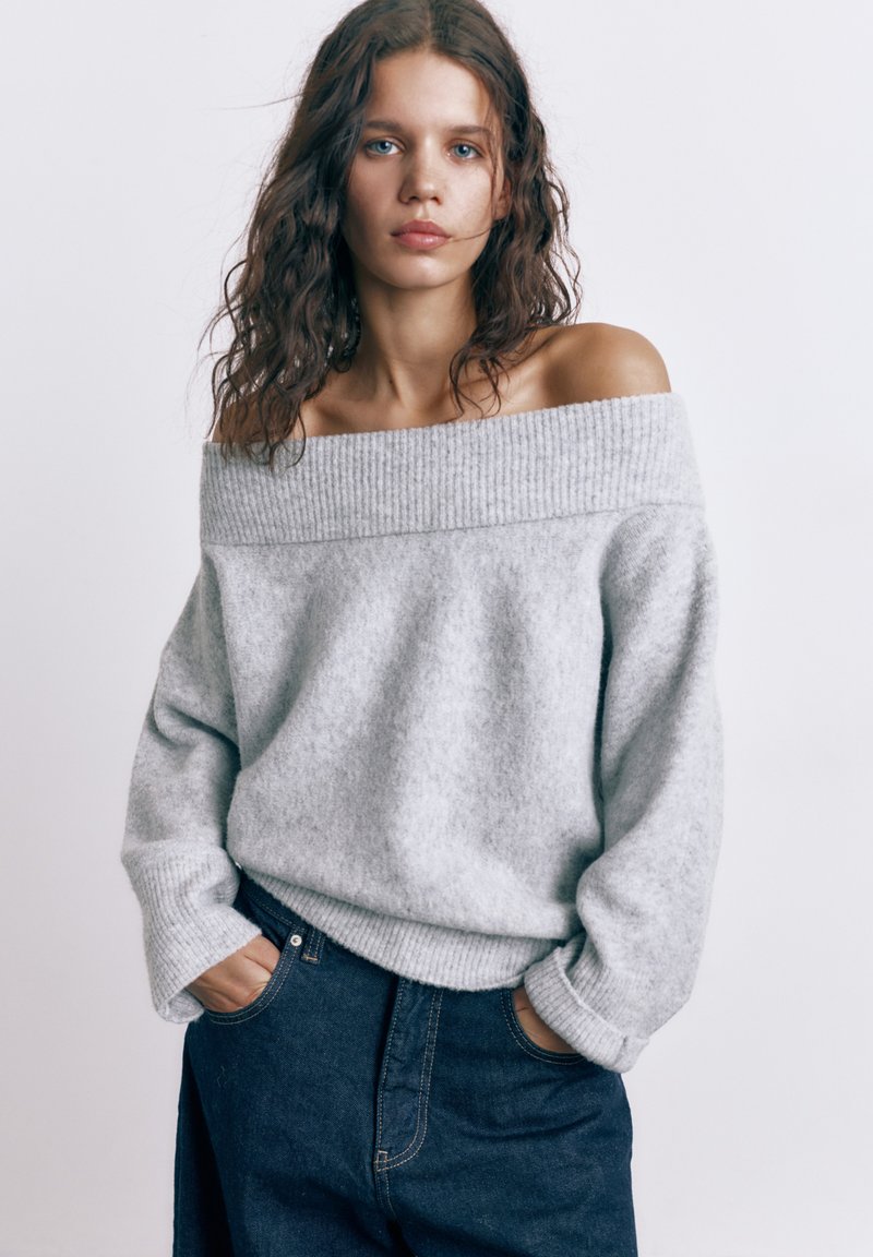 Mladá žena s kudrnatými vlasy, oblečená v šedém svetru se spuštěnými rameny a tmavě modrých džínách, s rukama v kapsách, čelem k přední straně na jednobarevném pozadí.