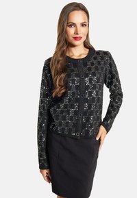 Cardigan nero con un motivo a scacchi di paillettes, collo rotondo, chiusura con bottoni e maniche lunghe. Abbinato a una gonna a tubino nera.