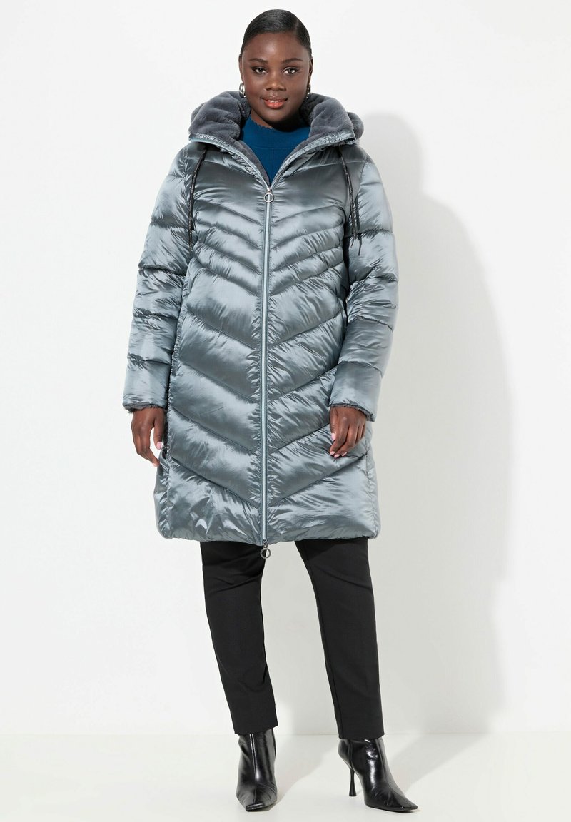Cappotto trapuntato grigio chiaro con design a zigzag, dotato di cappuccio foderato in pelliccia, cerniera frontale e tasche laterali, indossato sopra pantaloni neri e tacchi.
