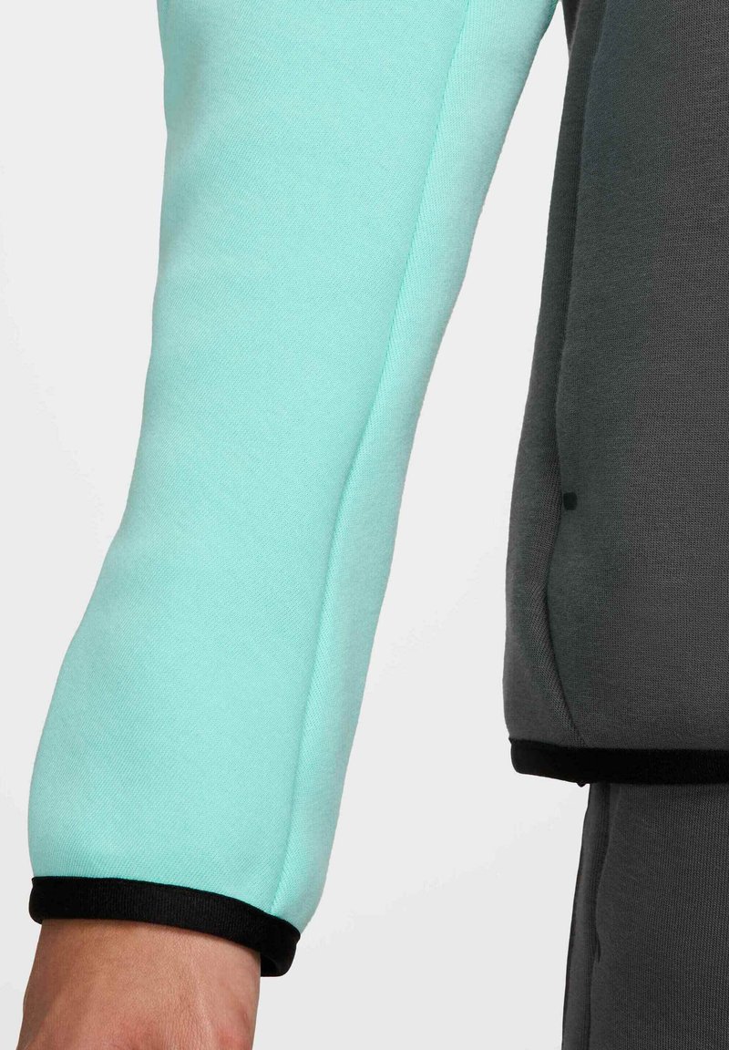 Top de manga larga que presenta una tela verde menta de textura suave con una sección en gris oscuro de contraste, terminado con puños negros.