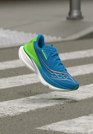 Chaussure de course bleue et verte en plein air au-dessus d’un passage piéton sur une route asphaltée, mettant en avant sa semelle blanche épaisse et son design aérodynamique.