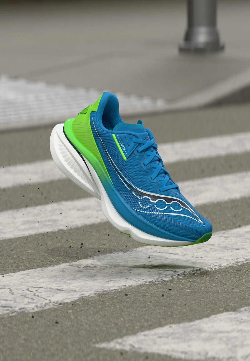 Μπλε και πράσινο αθλητικό παπούτσι στον αέρα πάνω από διάβαση πεζών σε ασφάλτινο δρόμο, παρουσιάζοντας την παχιά λευκή σόλα και τον αεροδυναμικό του σχεδιασμό.