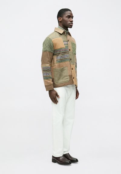 Casaco de camisa com múltiplos padrões em tons terrosos, apresentando remendos texturizados, botões e um grande bolso frontal. Combinado com calças brancas.