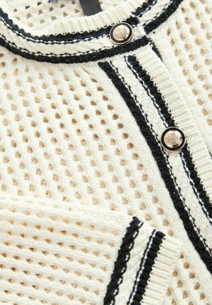 Knitted cardigan in cream with a textured mesh pattern, featuring black trim and buttons for closure. Notable striped detailing along edges.