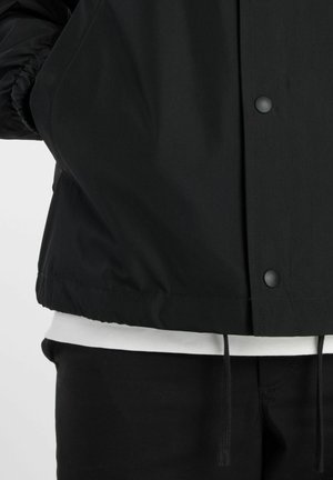 Giacca nera con bottoni a pressione sopra camicia bianca e pantaloni neri, che mostra l'orlo della giacca e i dettagli del cordino.