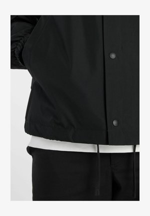 Giacca nera con bottoni a pressione sopra camicia bianca e pantaloni neri, che mostra l'orlo della giacca e i dettagli del cordino.