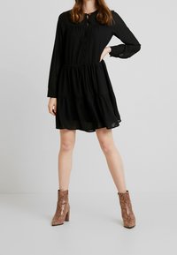 Černé šaty s dlouhým rukávem a tiered designem, se zapínáním na knoflíky v přední části, doplněné texturovanými hnědými kotníkovými botami s hadím vzorem.